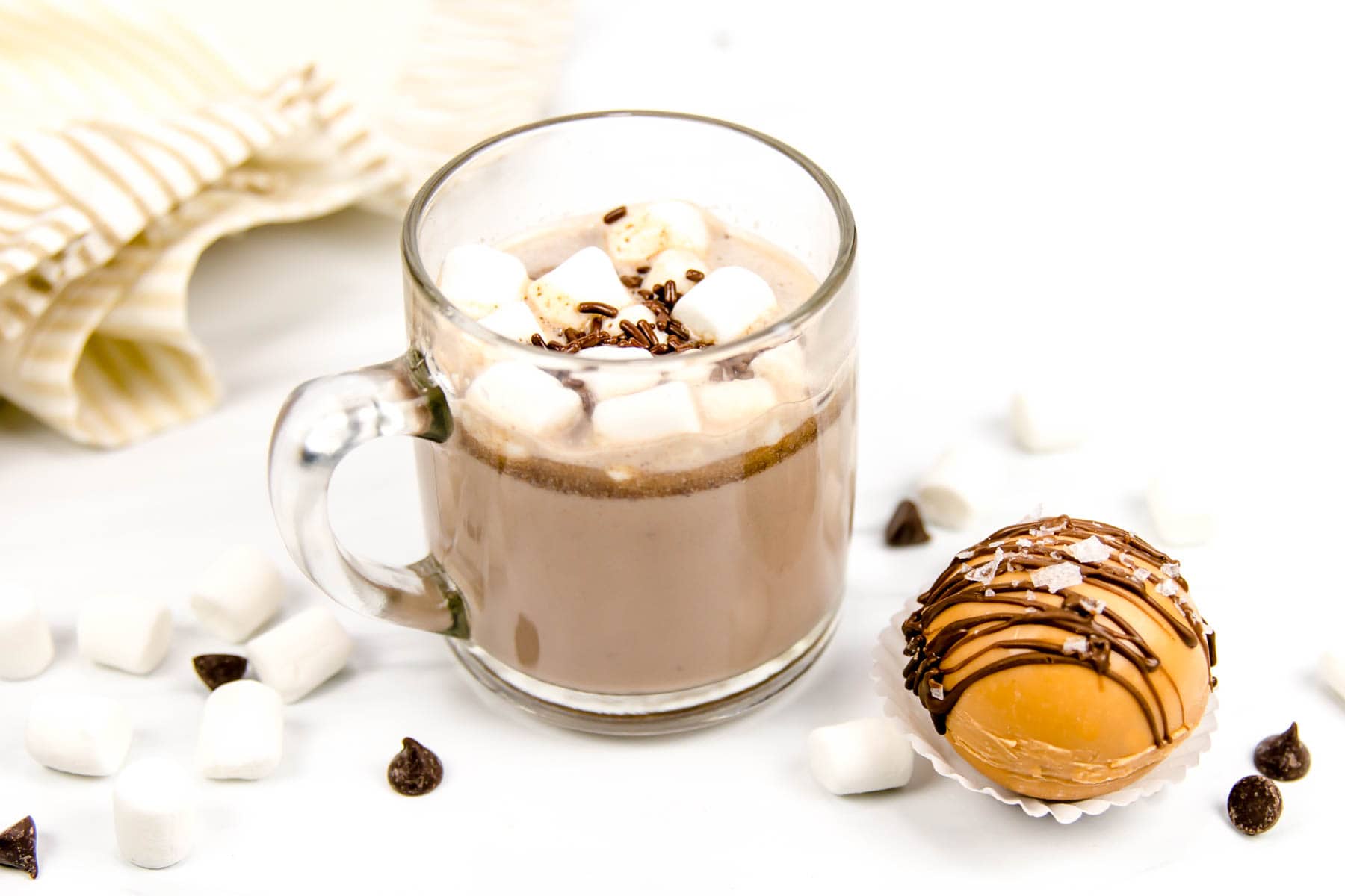 A clear glass mug filled with hot chocolate topped with mini marshmallows and chocolate sprinkles sits beside a round, chocolate-drizzled treat on a white surface.