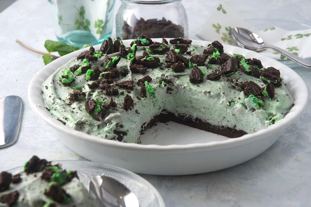A mint Oreo pie with a slice missing, topped with crushed chocolate cookies and green mint pieces, in a white dish on a light surface with utensils and napkin nearby.