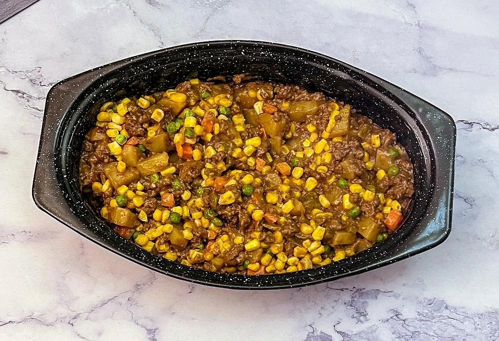 A black oval dish filled with a mixture of ground beef, corn, peas, carrots, and diced potatoes in a brown sauce, placed on a marble surface.