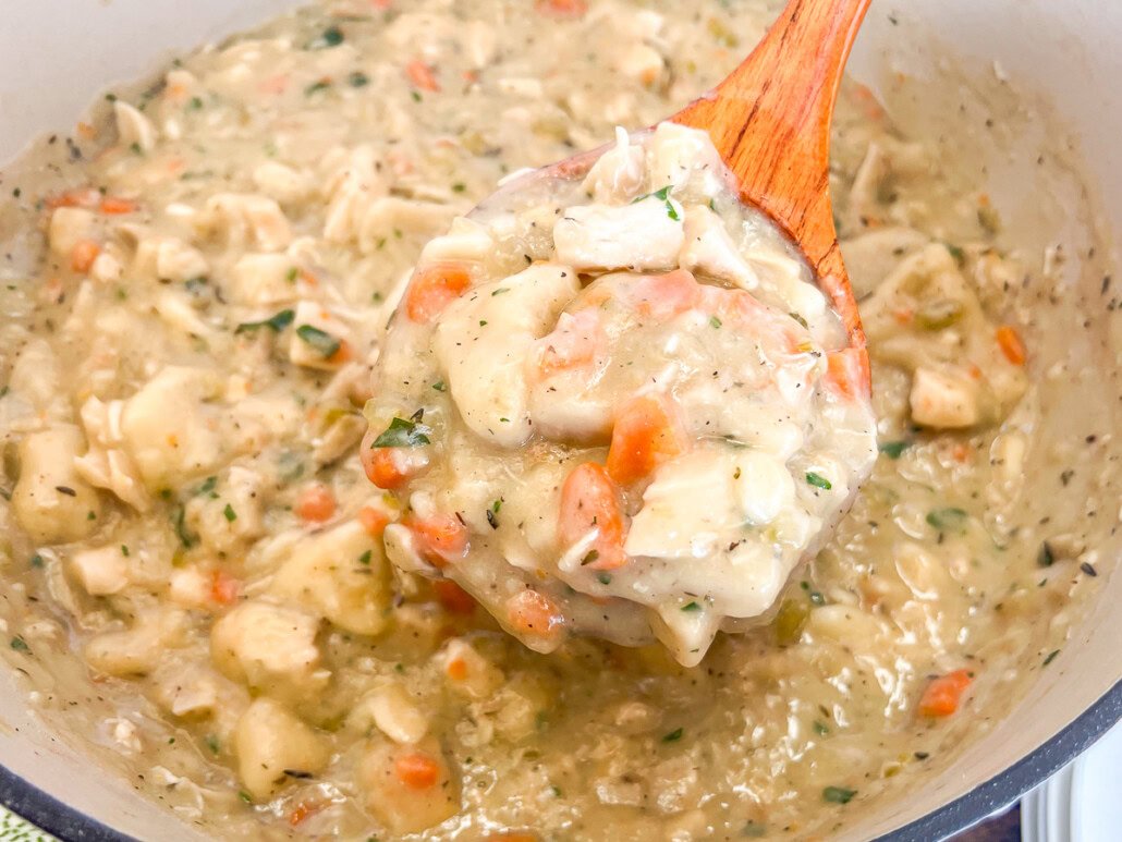 A wooden spoon lifts a portion of thick, creamy chicken and dumplings with carrots and herbs from a pot.