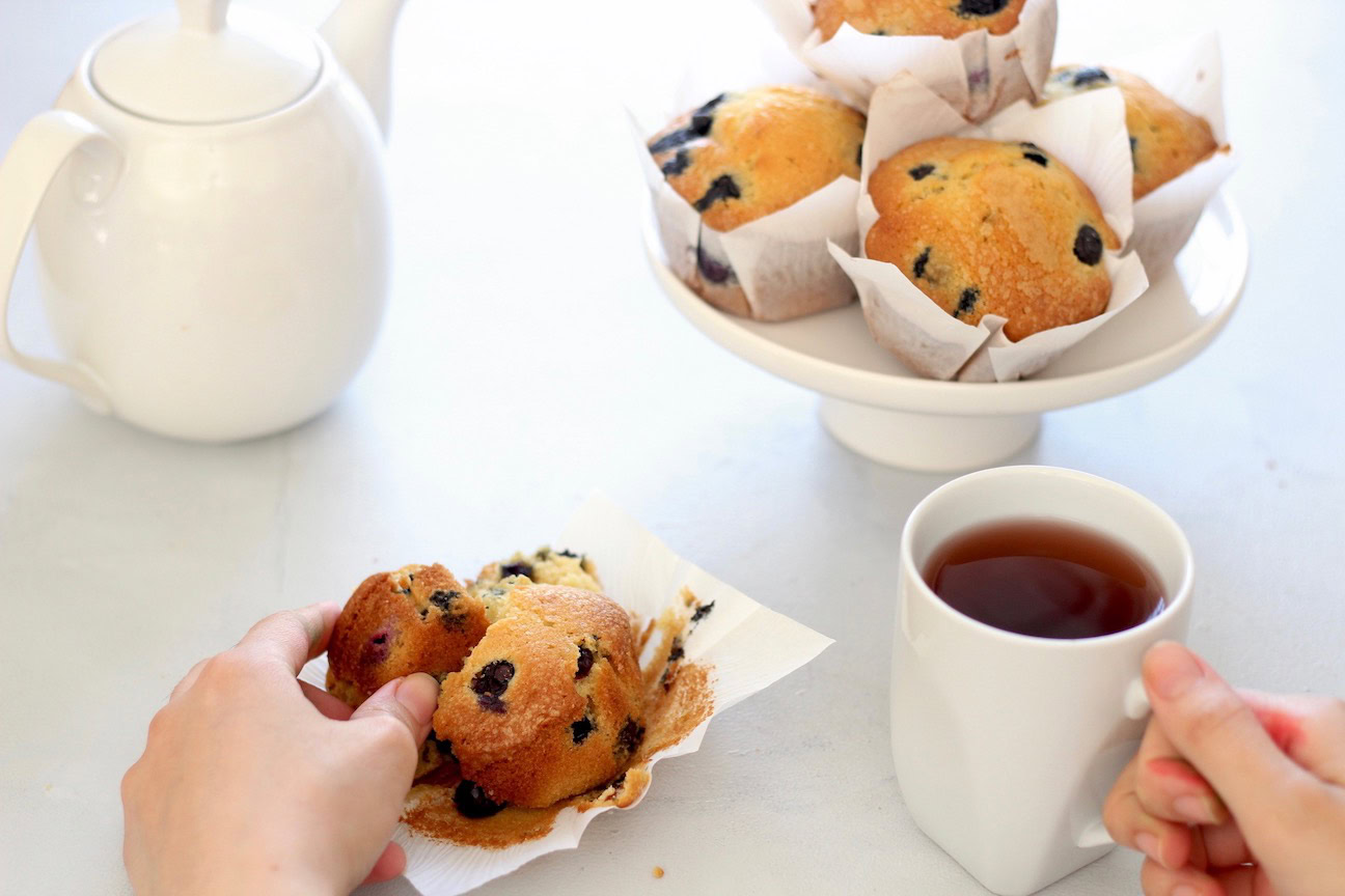 A person breaks apart a blueberry muffin on a napkin while another person holds a cup of tea; more muffins and a teapot are also on the table.