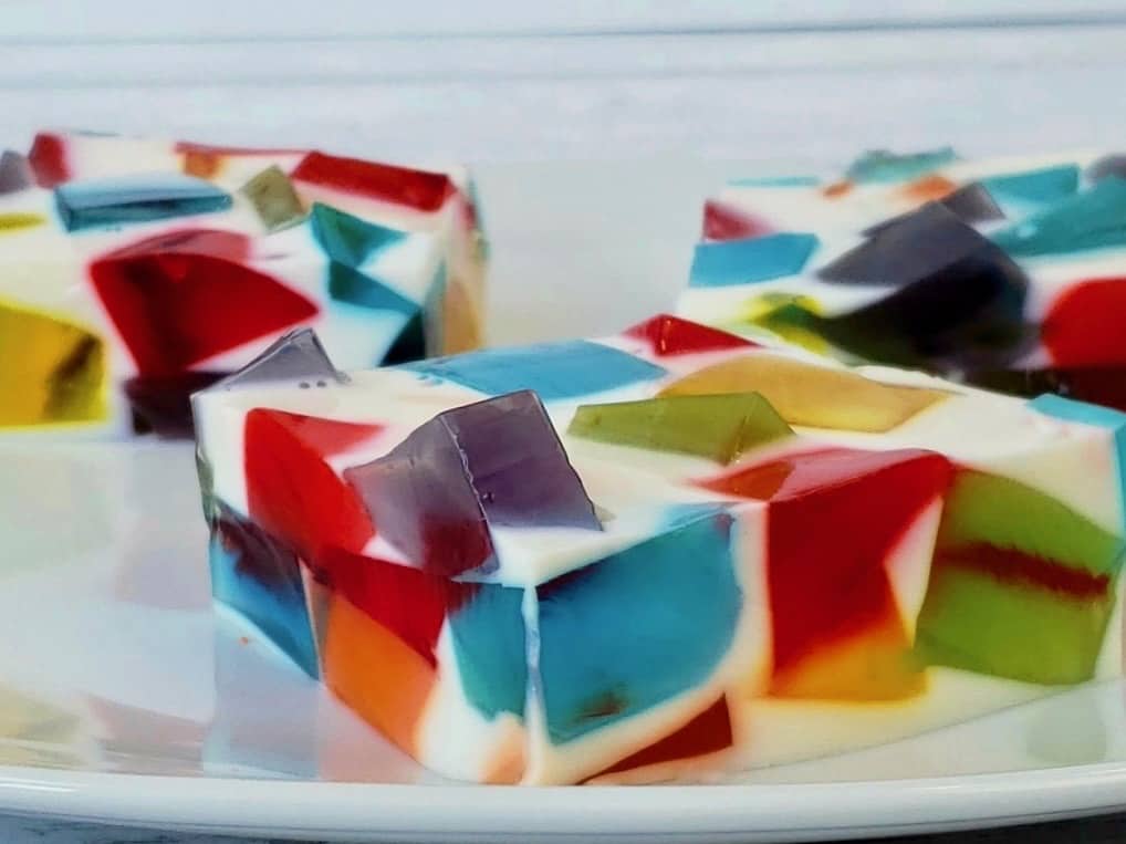 A close-up of colorful mosaic gelatin dessert squares on a white plate, showing embedded cubes of red, blue, green, orange, and purple jelly in a white gelatin base.