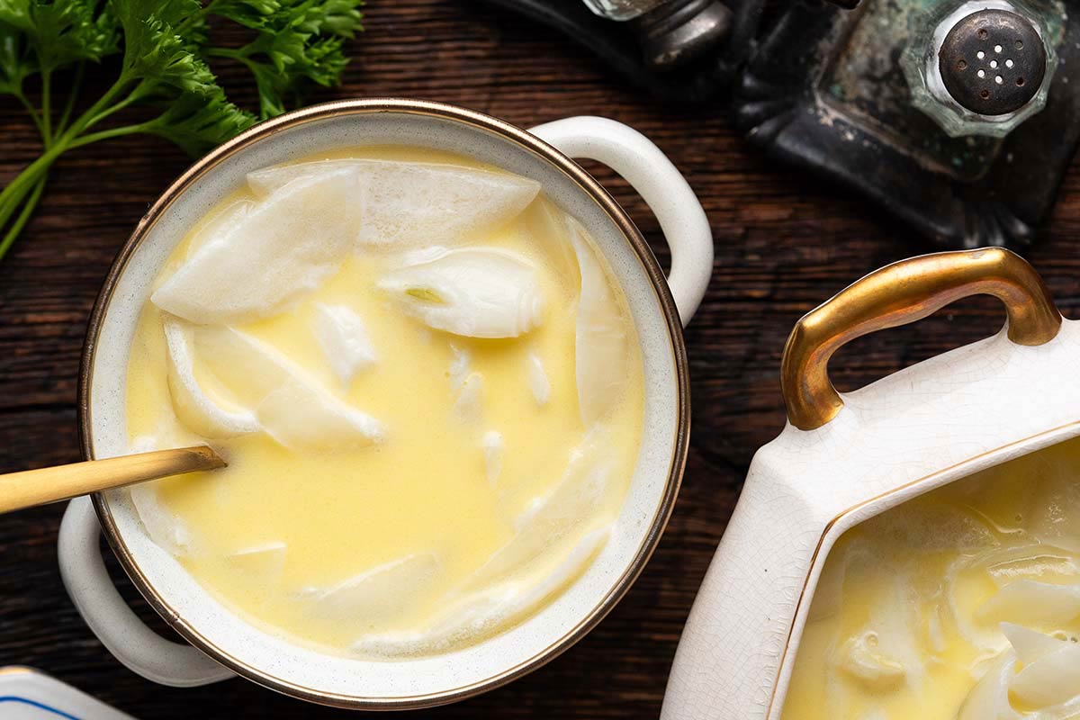 A bowl of creamy yellow soup with visible onion slices, a gold spoon, and a sprig of parsley on a dark wooden table.