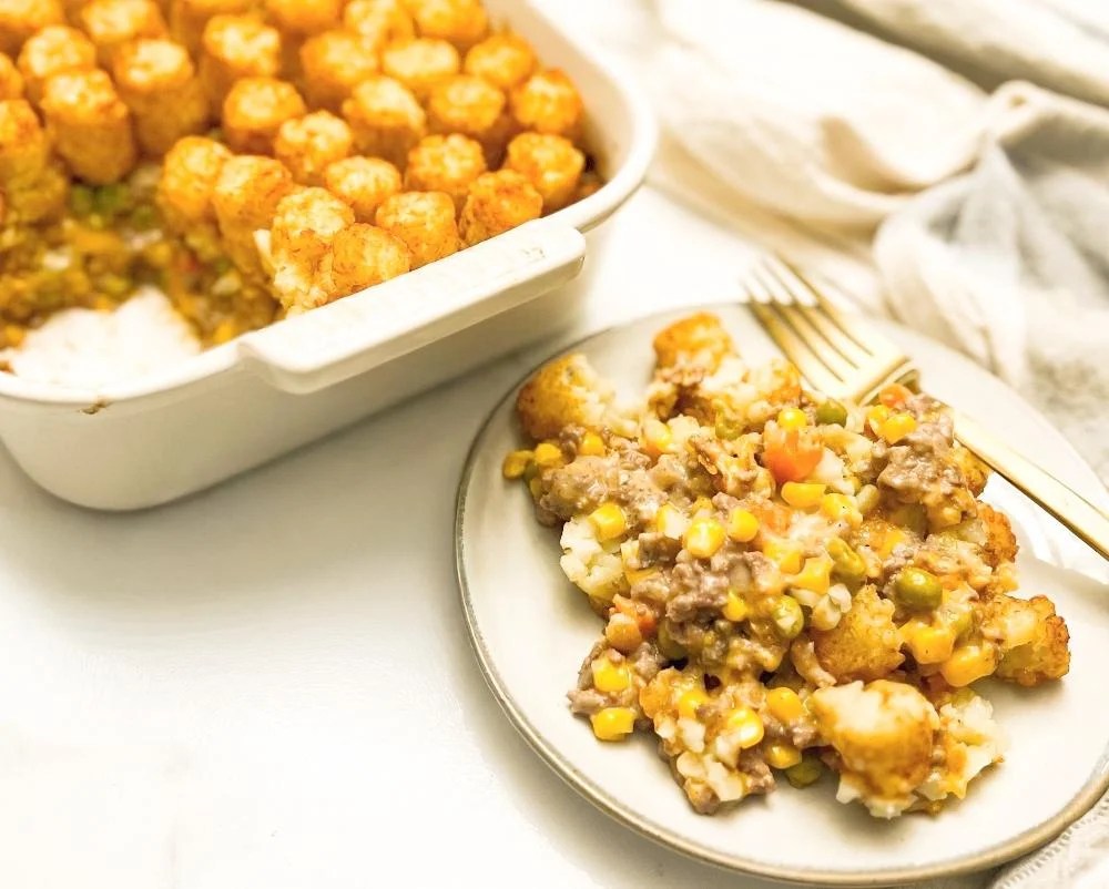 A plate of tater tot casserole with ground beef, mixed vegetables, and corn sits beside a baking dish of the same casserole on a white surface.