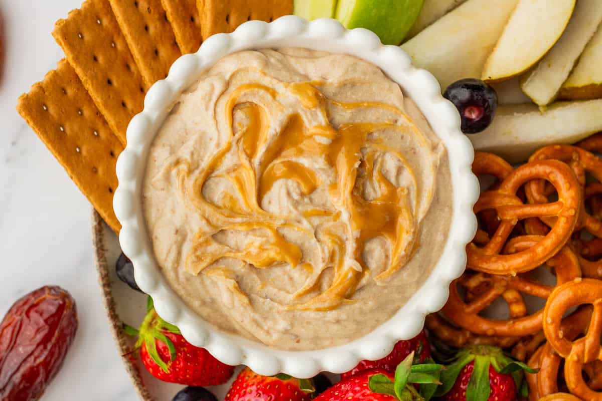 A white bowl of creamy dip with caramel swirls, surrounded by crackers, pretzels, strawberries, apple and pear slices, and dates.