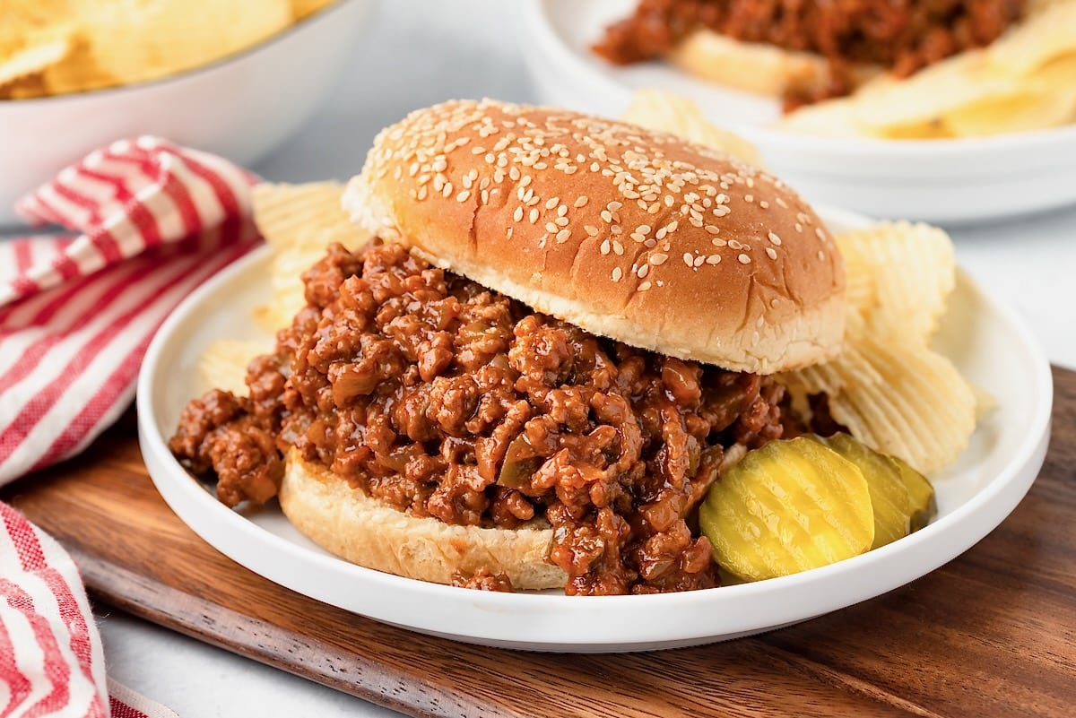 A sloppy joe sandwich on a sesame bun is served on a white plate with potato chips and pickle slices, next to a red and white striped napkin.