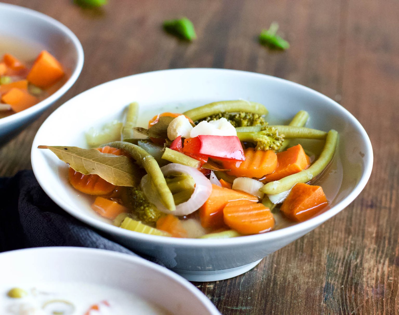 A bowl of vegetable soup containing carrots, green beans, broccoli, celery, and garnished with sliced radishes.