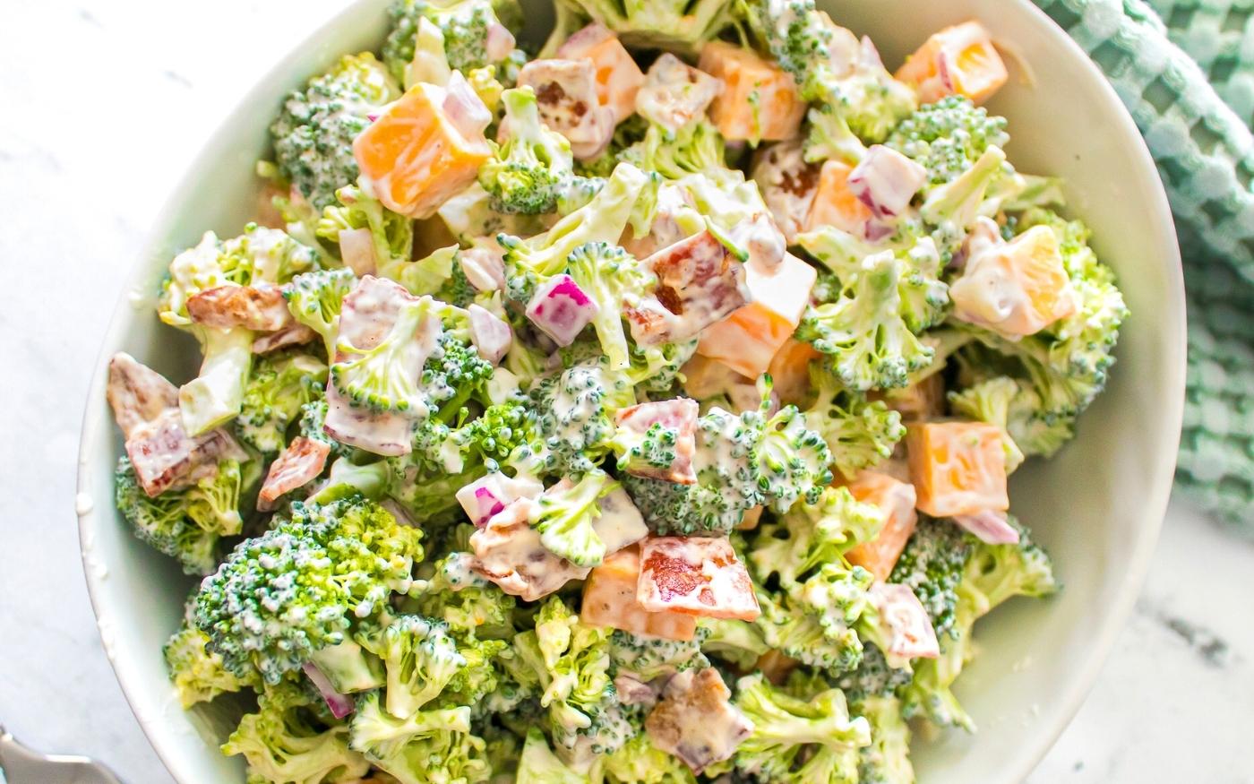 A bowl of broccoli salad mixed with diced cheddar cheese, red onions, bacon pieces, and a creamy dressing.