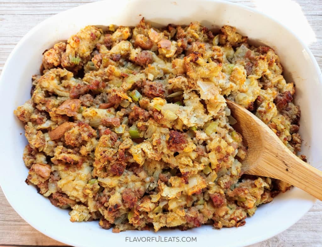 A casserole dish filled with baked stuffing made of bread, sausage, and vegetables, with a wooden spoon resting inside.