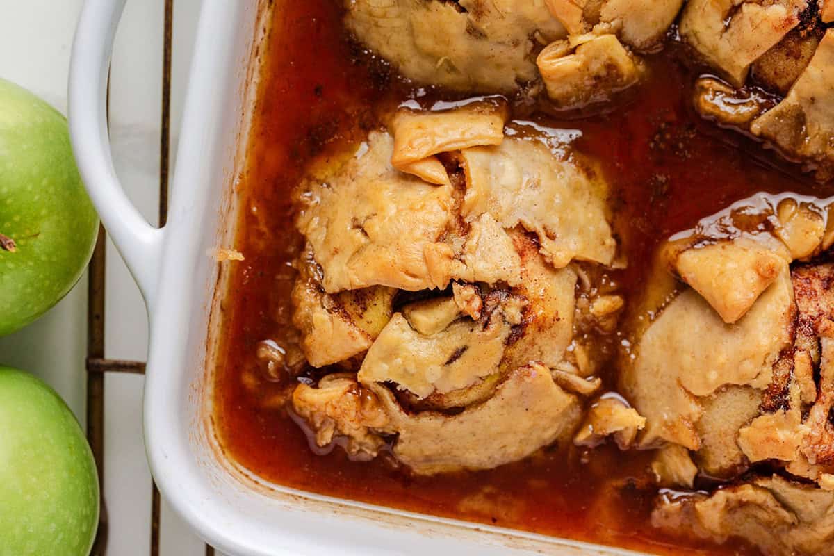Close-up of baked apple dumplings in a caramelized sauce inside a white baking dish, with green apples beside the dish.