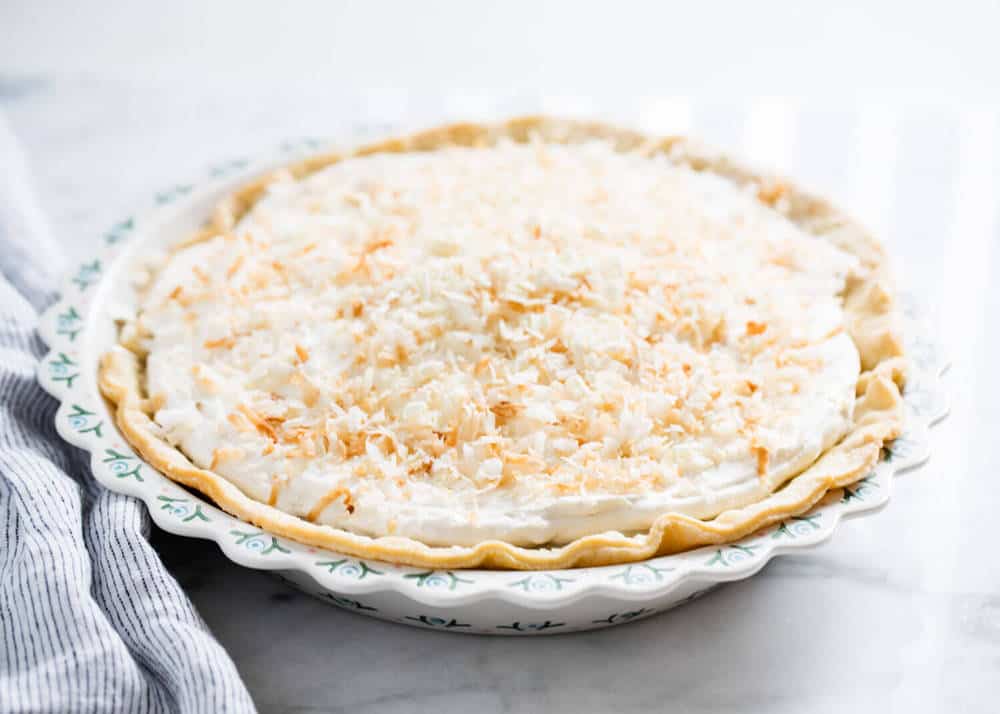 A coconut cream pie with a flaky crust, topped with whipped cream and toasted coconut, sits on a patterned pie plate on a marble surface.