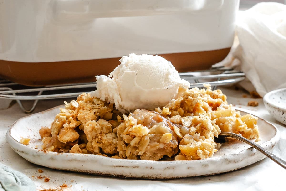 A serving of apple crisp topped with a scoop of vanilla ice cream on a ceramic plate with a fork beside it.