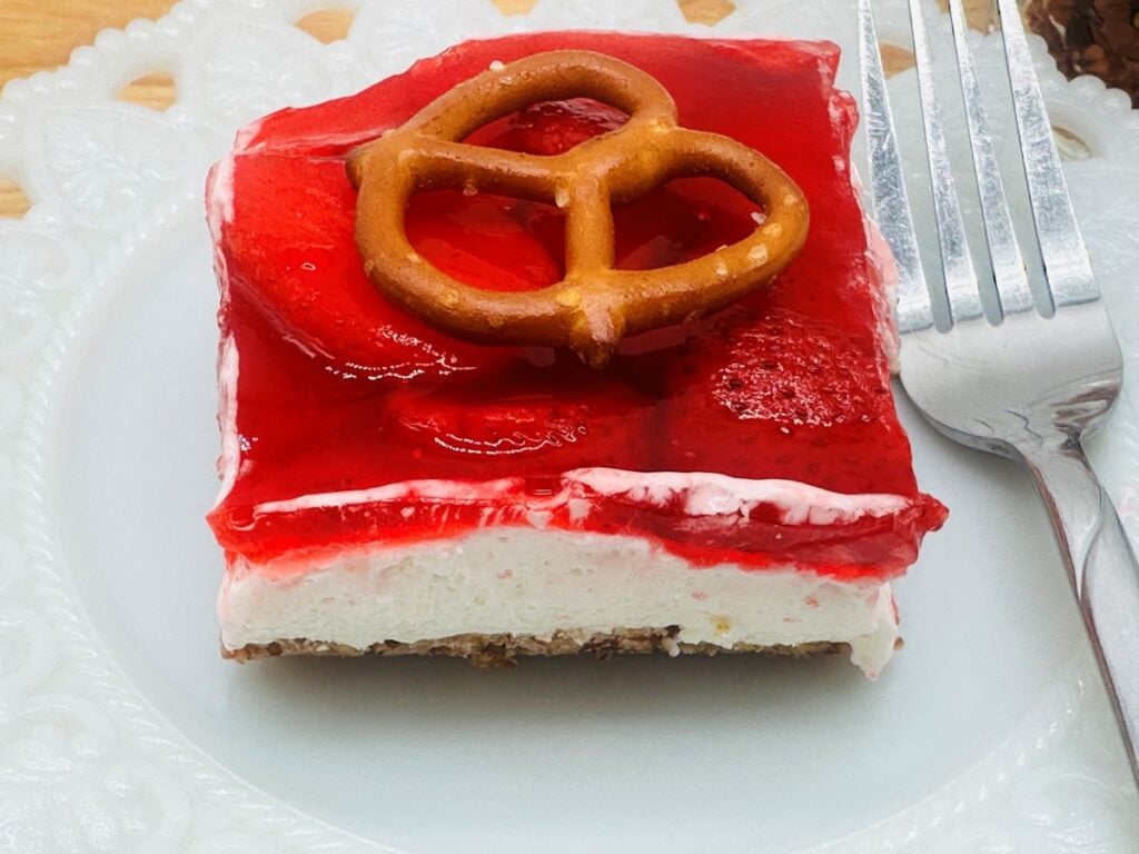 A square slice of strawberry pretzel dessert with a pretzel on top, layered with red gelatin and cream, served on a white plate with a fork.