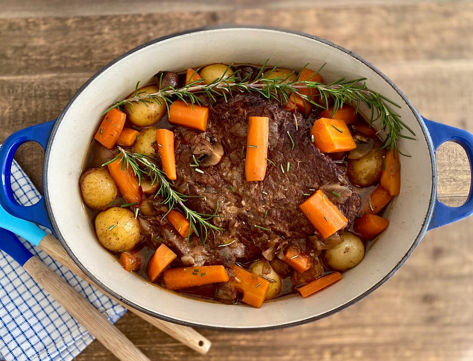 A cooked beef pot roast with potatoes, carrots, mushrooms, and sprigs of rosemary in a blue Dutch oven on a wooden table.