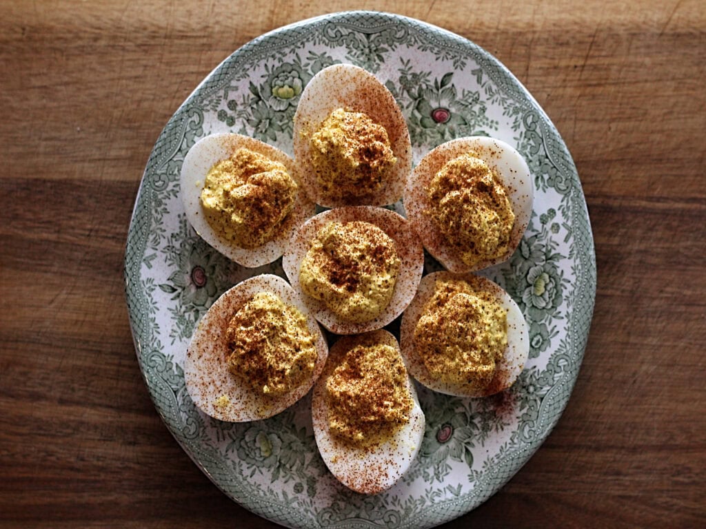A plate of six deviled eggs topped with paprika sits on a floral-patterned dish on a wooden surface.