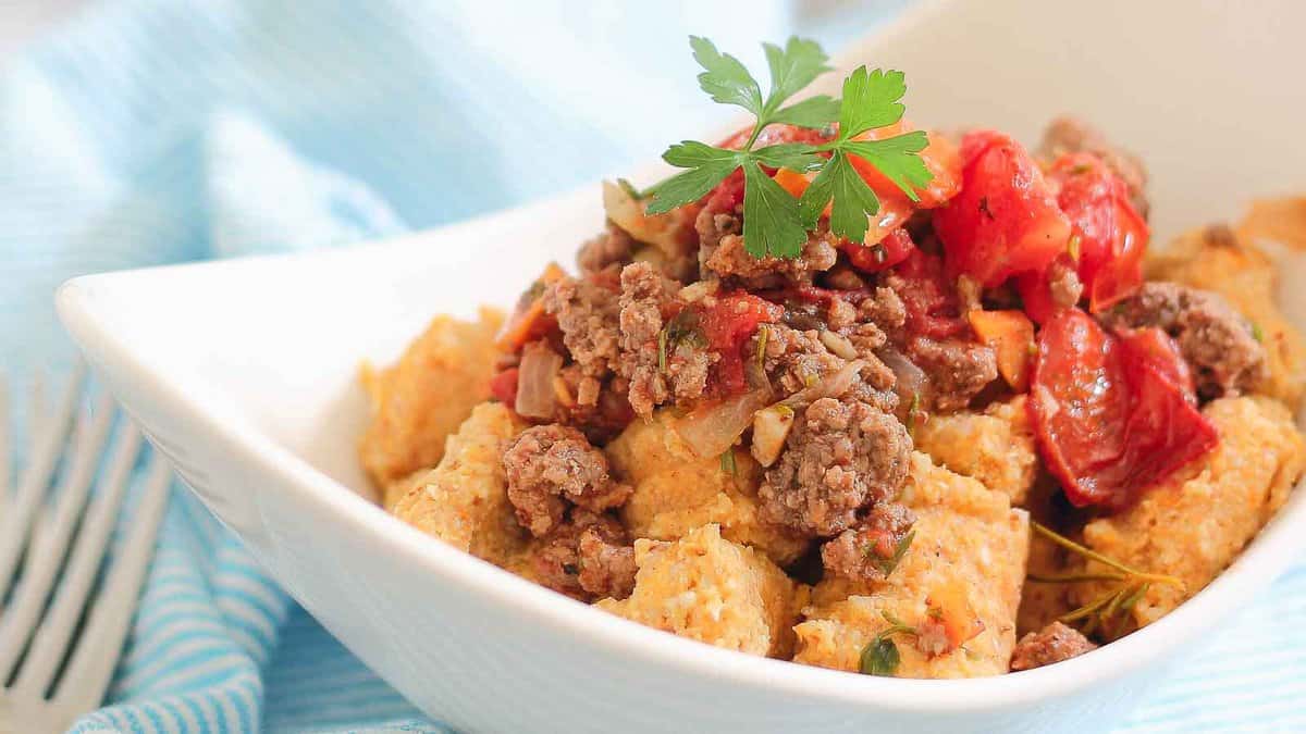 A white bowl filled with ground beef, tomatoes, onions, and cubes of cornbread, garnished with a sprig of parsley.