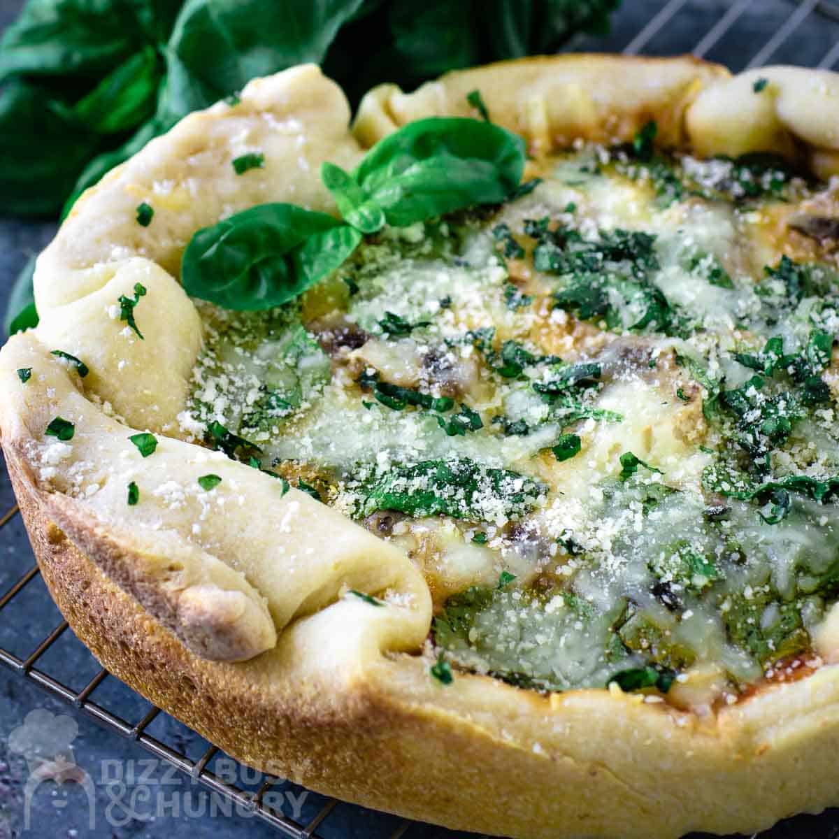 A deep-dish spinach and cheese pizza with a golden crust, topped with fresh basil leaves and grated parmesan, on a cooling rack.