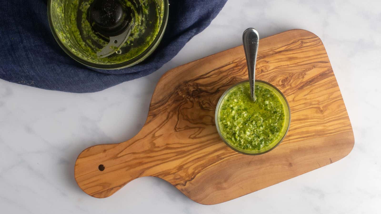 A glass bowl of green pesto with a spoon sits on a wooden cutting board; a partially filled food processor is nearby on a marble surface.