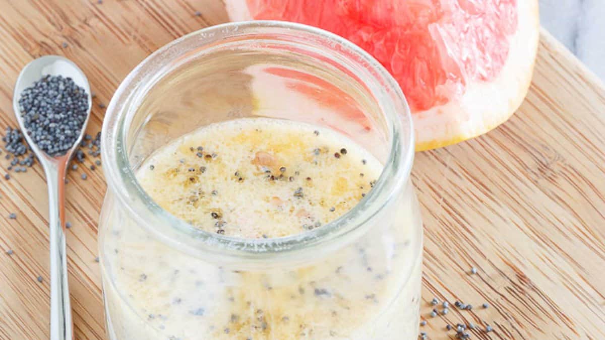 A glass jar of poppy seed salad dressing sits on a wooden surface with a spoonful of poppy seeds and a slice of grapefruit nearby.