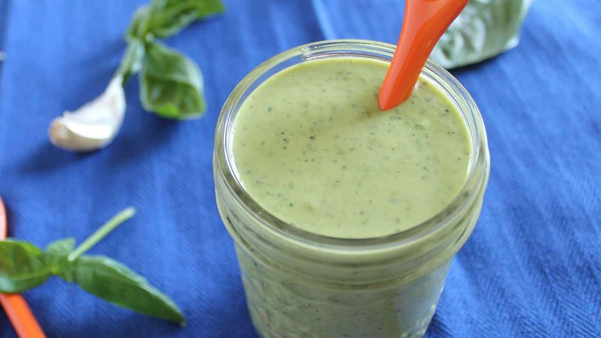 A jar of green creamy sauce with an orange spoon, placed on a blue cloth, with basil leaves and a garlic clove nearby.