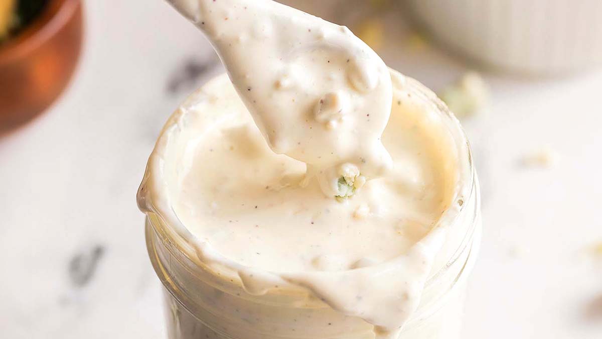 A spoon dipped in thick, creamy white dressing with visible chunks, held above a glass jar filled with the same dressing.