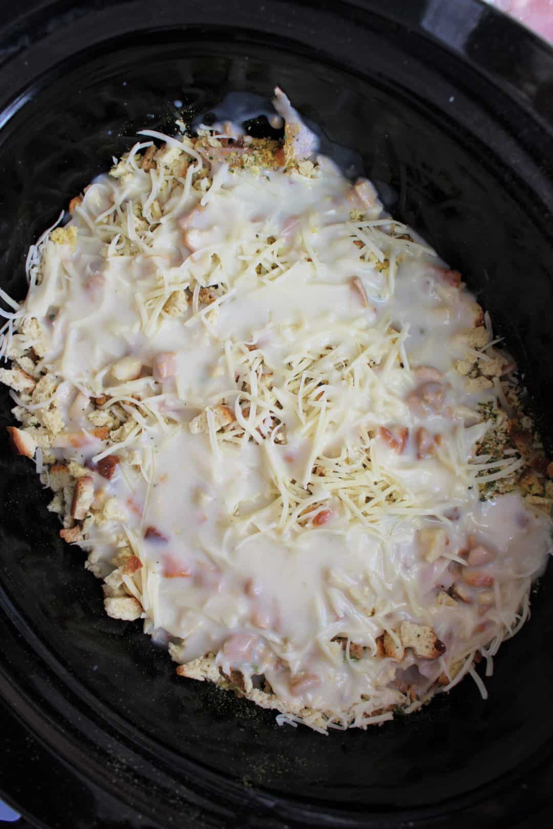 A slow cooker filled with cubed bread, shredded cheese, and creamy sauce, prepared for cooking.