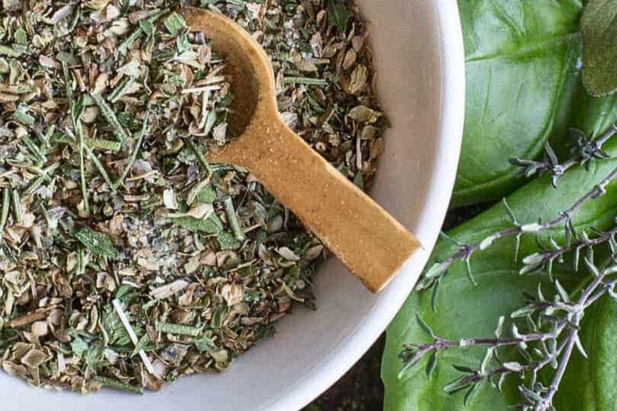 A white bowl filled with dried mixed herbs and a wooden spoon, next to fresh basil leaves and sprigs of fresh herbs.