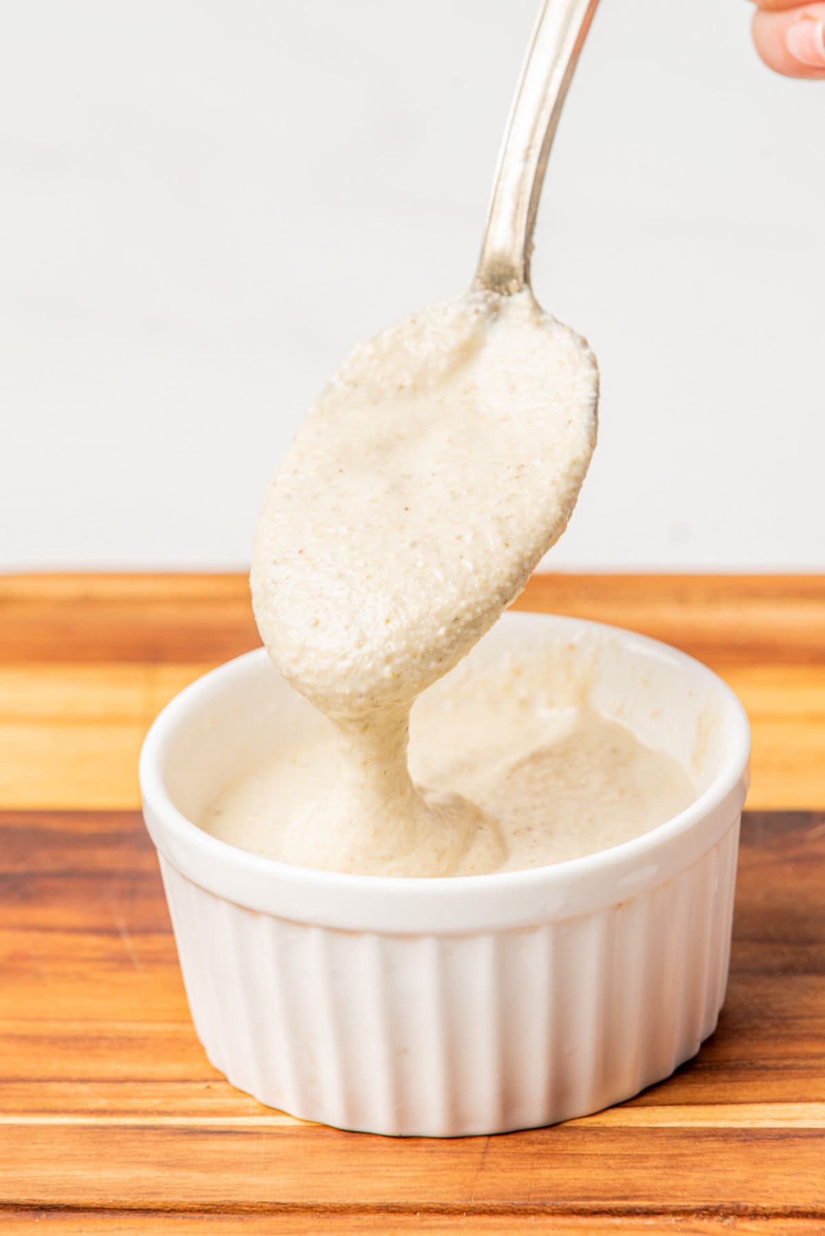 A spoon lifts a thick, creamy beige sauce from a small white ramekin placed on a wooden surface.