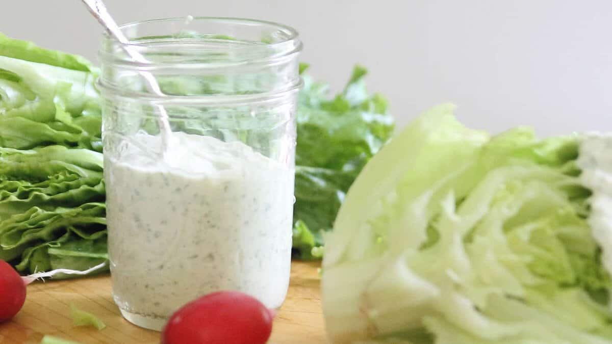 A glass jar filled with creamy ranch dressing sits on a wooden surface, surrounded by leafy lettuce and a red radish, with a silver spoon in the jar.