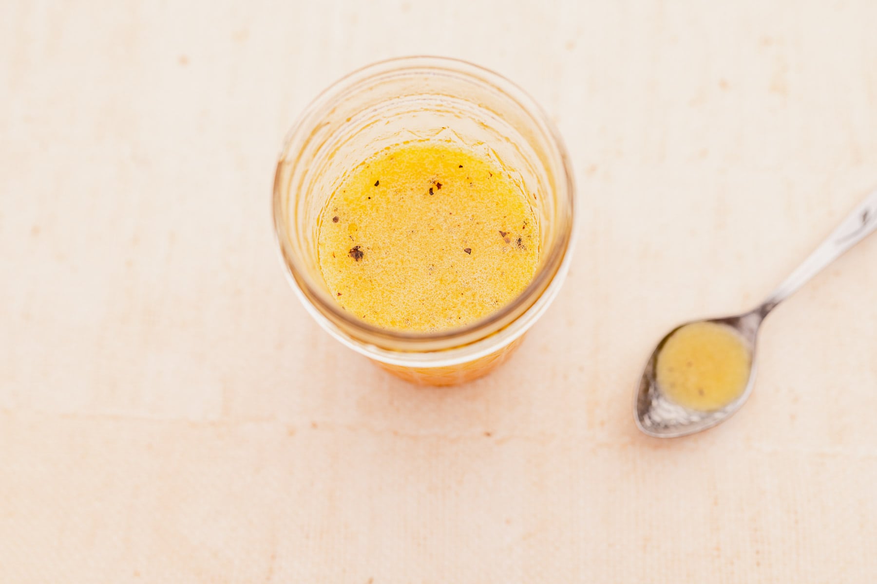 A glass jar containing a small amount of yellow liquid sits next to a metal spoon with the same liquid on a light-colored surface.