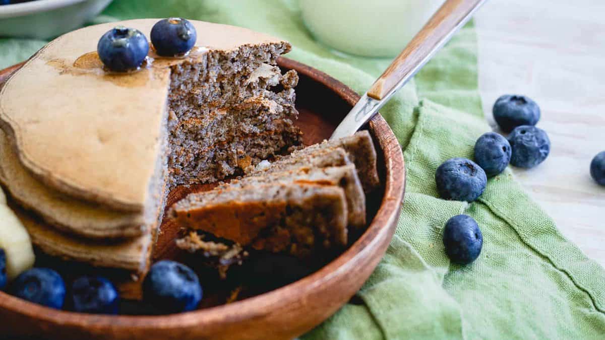 A stack of pancakes with blueberries on top, partially sliced, sits on a wooden plate with a knife. Extra blueberries are scattered around.