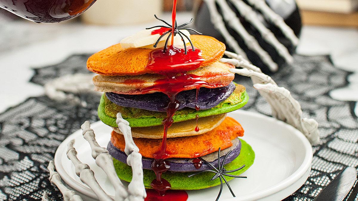 A stack of colorful pancakes with red syrup, topped with butter and plastic spiders, sits on a plate held by a skeleton hand.
