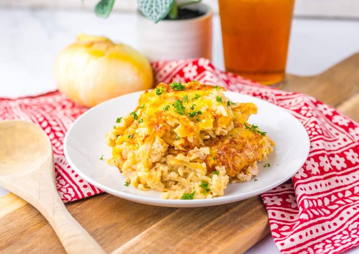 A plate of cheesy casserole with rice and herbs sits on a white plate atop a red patterned cloth, next to a wooden spoon, onion, and a glass of iced tea.