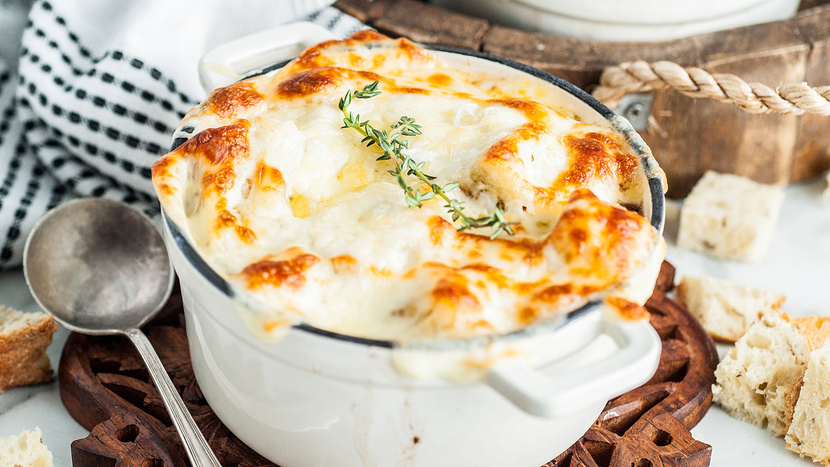 A white ceramic dish filled with baked French onion soup, topped with melted, browned cheese and garnished with fresh thyme. A spoon and pieces of bread are nearby.