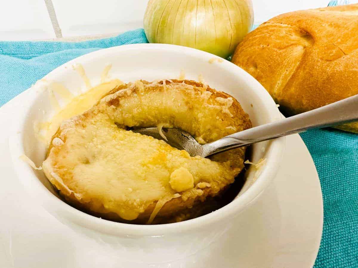 A bowl of French onion soup topped with melted cheese and bread, with a spoon inside. An onion and a bread roll are in the background on a blue cloth.