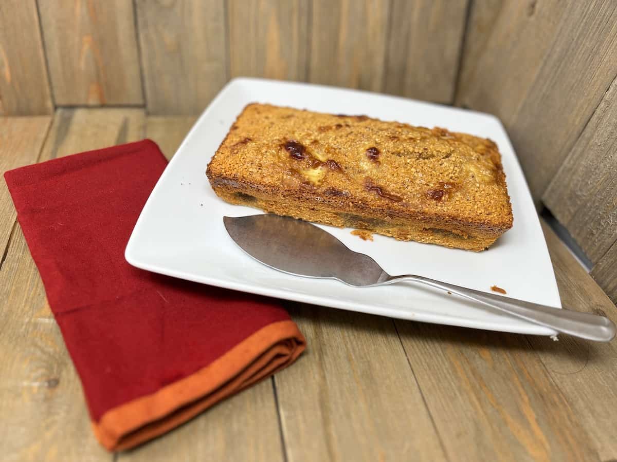 A rectangular loaf of bread or cake on a white square plate with a serving utensil, next to a folded red napkin on a wooden surface.