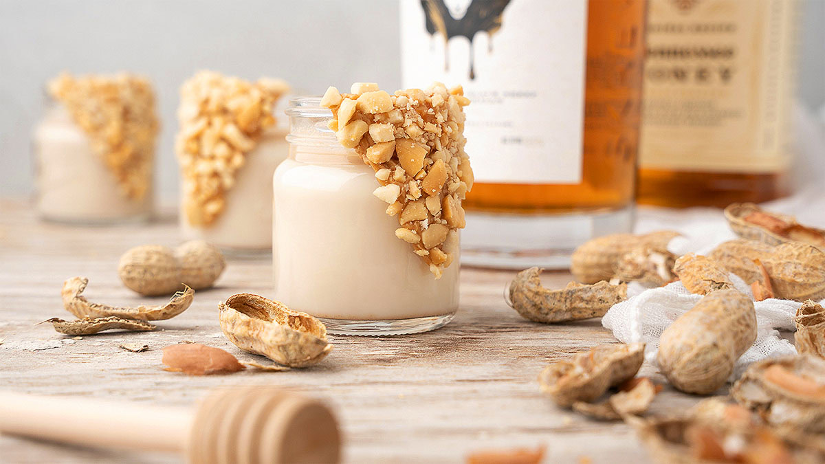 Small glass jar filled with creamy white dessert, topped with chopped nuts, surrounded by peanut shells and honey dipper, with bottles in the background.