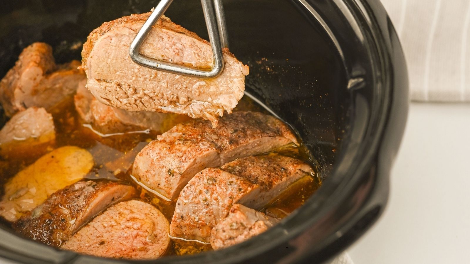 Sliced, cooked beef is shown in a slow cooker with one piece being lifted using metal tongs. The meat appears juicy and seasoned.