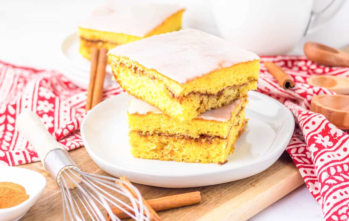 Two pieces of yellow cake with a cinnamon layer and white icing are stacked on a white plate, surrounded by baking utensils and a red patterned cloth.
