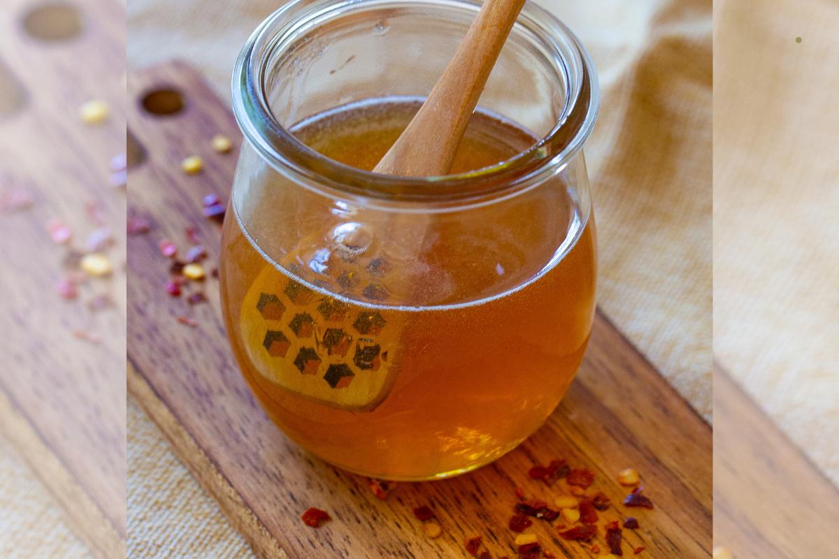A glass jar of honey with a wooden honey dipper inside, placed on a wooden board with scattered red pepper flakes nearby.