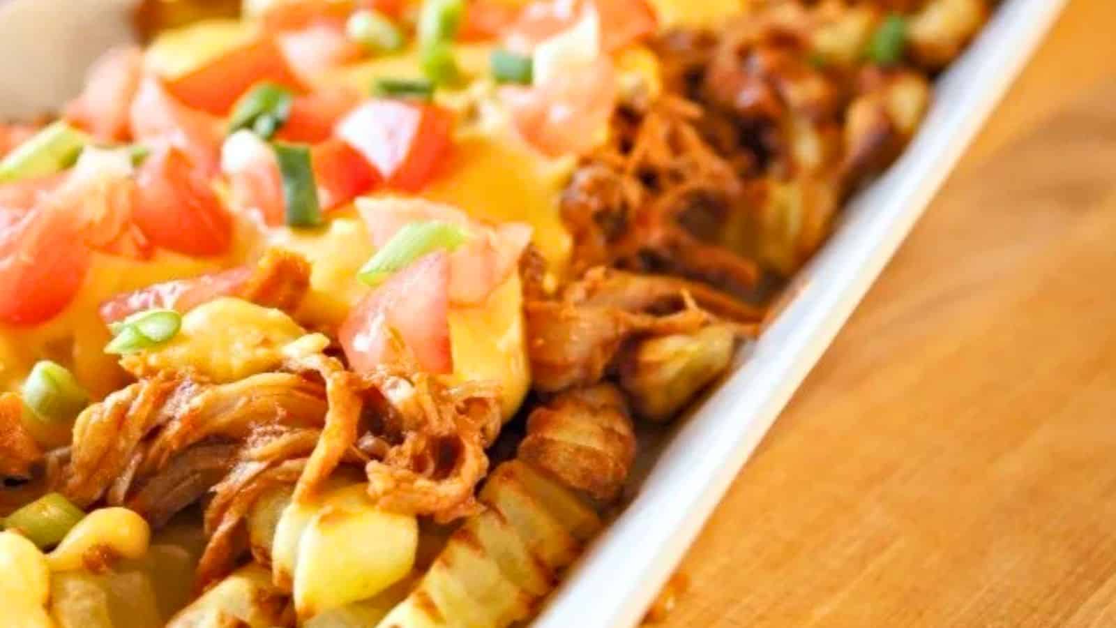 Crinkle-cut fries topped with shredded meat, melted cheese, diced tomatoes, and chopped green onions in a white dish on a wooden surface.