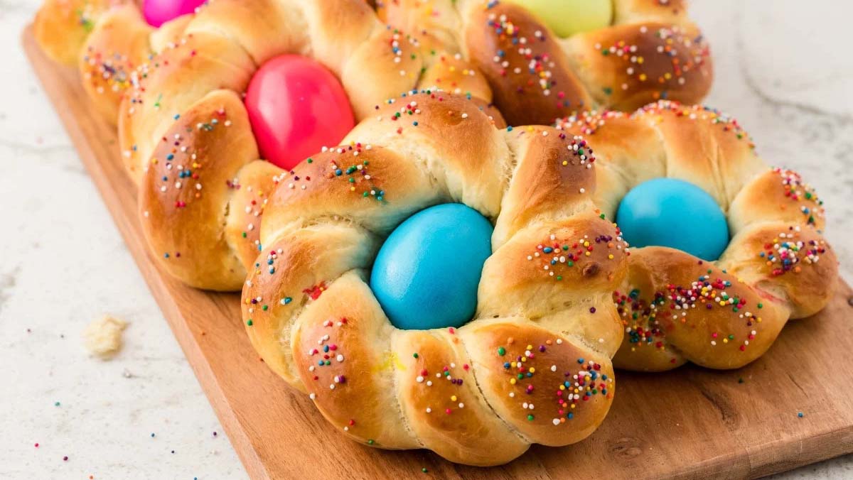 Braided sweet bread rings with colorful dyed eggs in the center, topped with rainbow sprinkles, displayed on a wooden board.