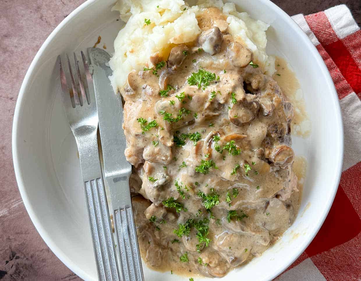 A plate of mashed potatoes topped with beef and mushroom gravy, garnished with chopped parsley, with a fork and knife on the side.