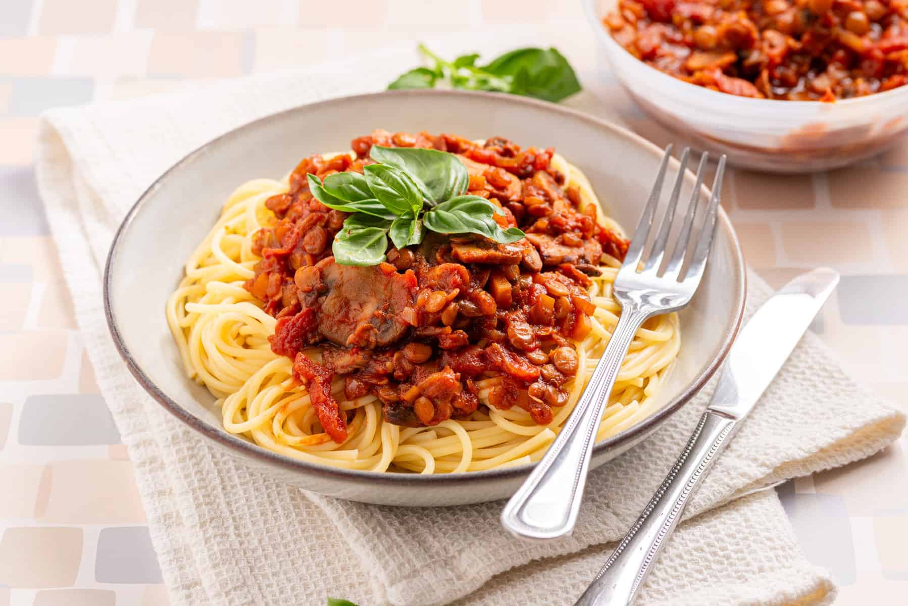 A plate of spaghetti topped with tomato-based lentil and vegetable sauce, garnished with fresh basil, with a fork and knife on the side.