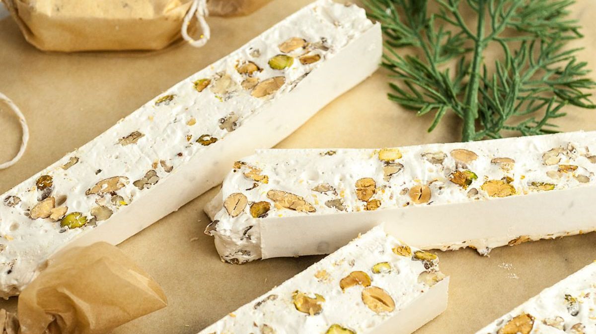 Three rectangular pieces of nougat with visible nuts are placed on parchment paper, next to a sprig of greenery and a partially wrapped candy.