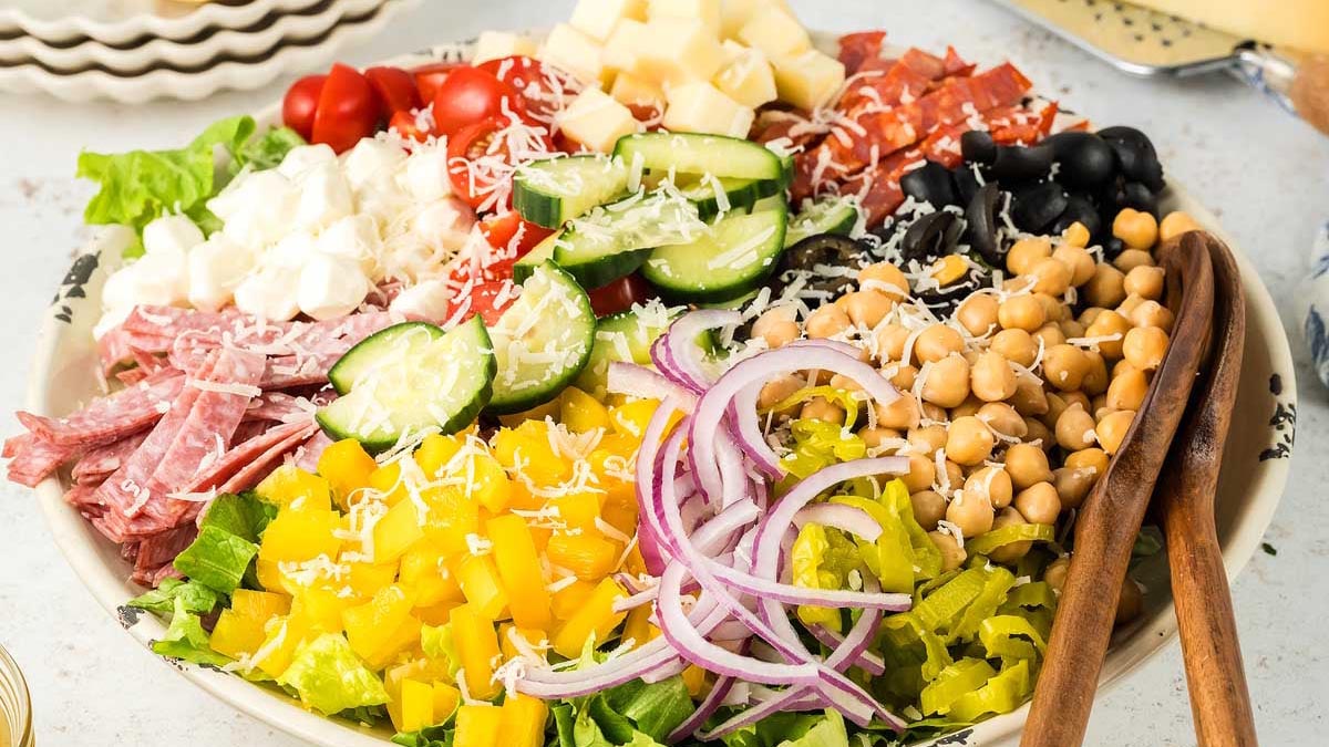 A large salad plate with chopped lettuce, yellow bell peppers, red onions, cucumbers, cherry tomatoes, chickpeas, cheese cubes, salami, olives, and shredded cheese, with wooden salad tongs.