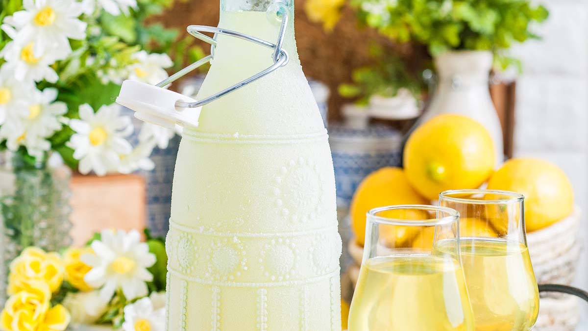 A decorative glass bottle filled with pale yellow liquid sits beside two glasses, with lemons and flowers in the background.