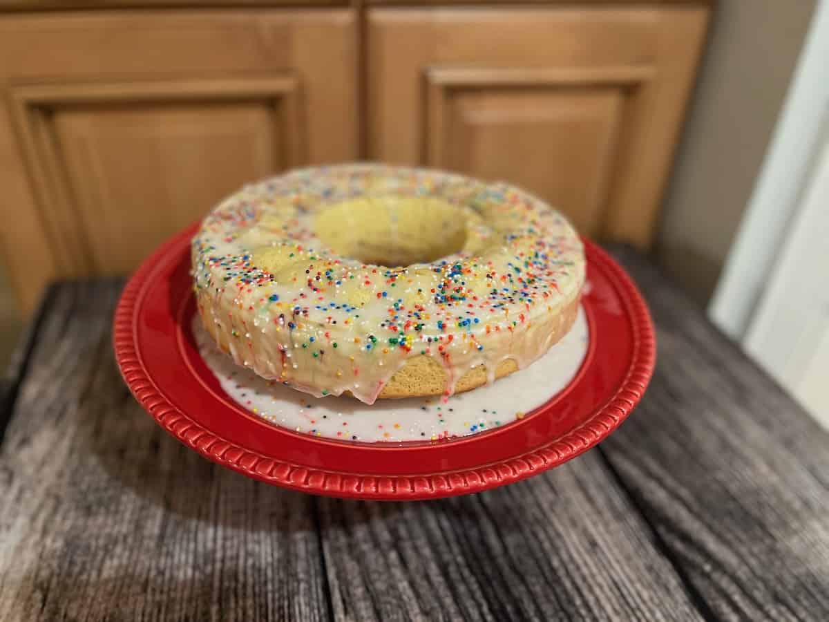 A round bundt cake with white icing and colorful sprinkles sits on a red cake stand on a wooden surface, with wooden cabinets in the background.