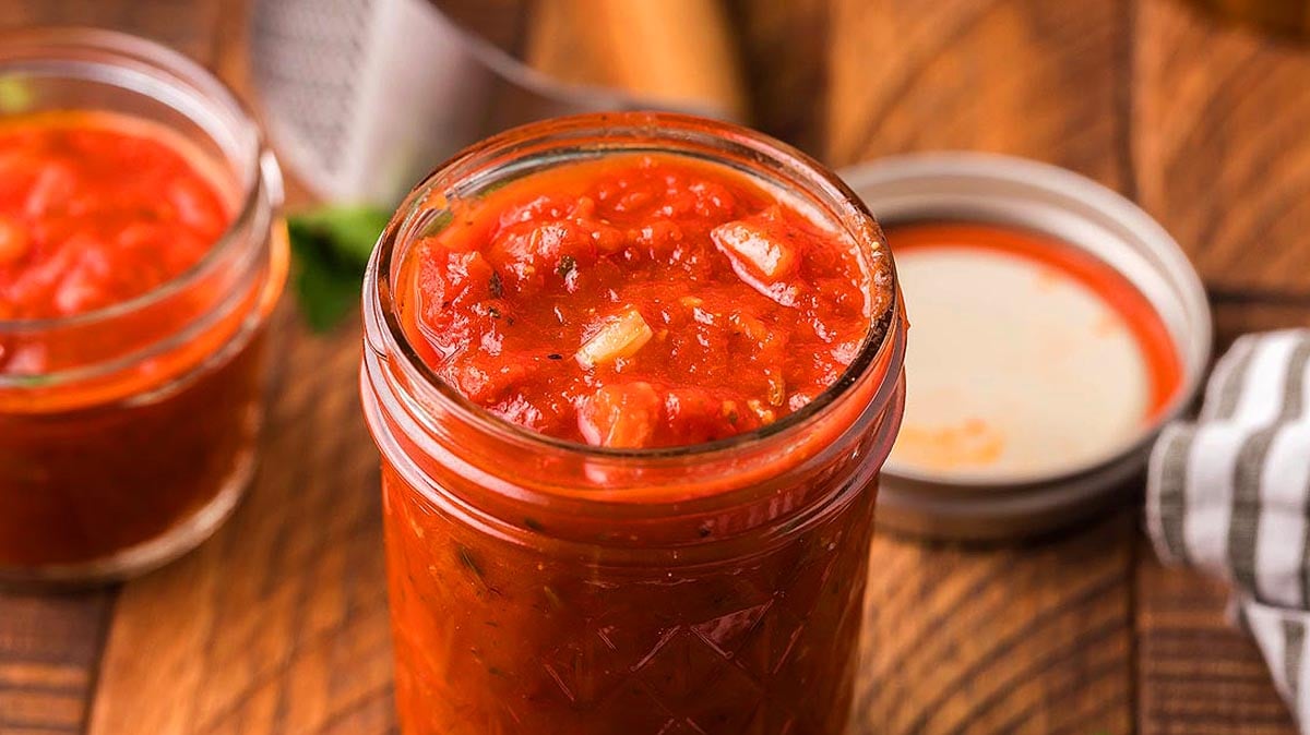 A glass jar filled with homemade tomato salsa sits on a wooden surface, with its lid off and another jar and kitchen towel visible in the background.