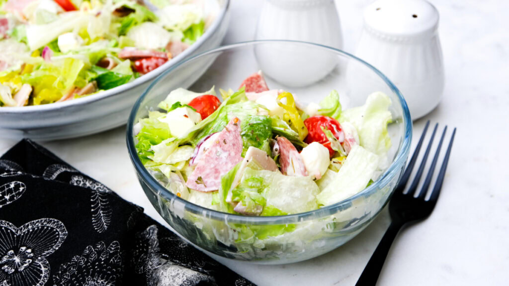 A clear glass bowl filled with a salad of lettuce, tomatoes, cheese, and sliced deli meat sits next to a black fork and two white shakers on a white surface.